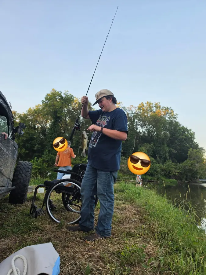 A man holding a fish beside Yadkin River, with a bicycle, a vehicle, and two companions nearby, enjoying great fishing and fun at Hillbilly Campground.