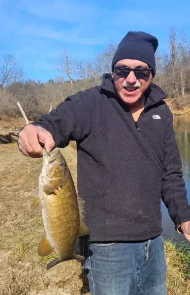 A smiling camper in sunglasses and a black knit cap holding a large bass caught from Fishing Creek.