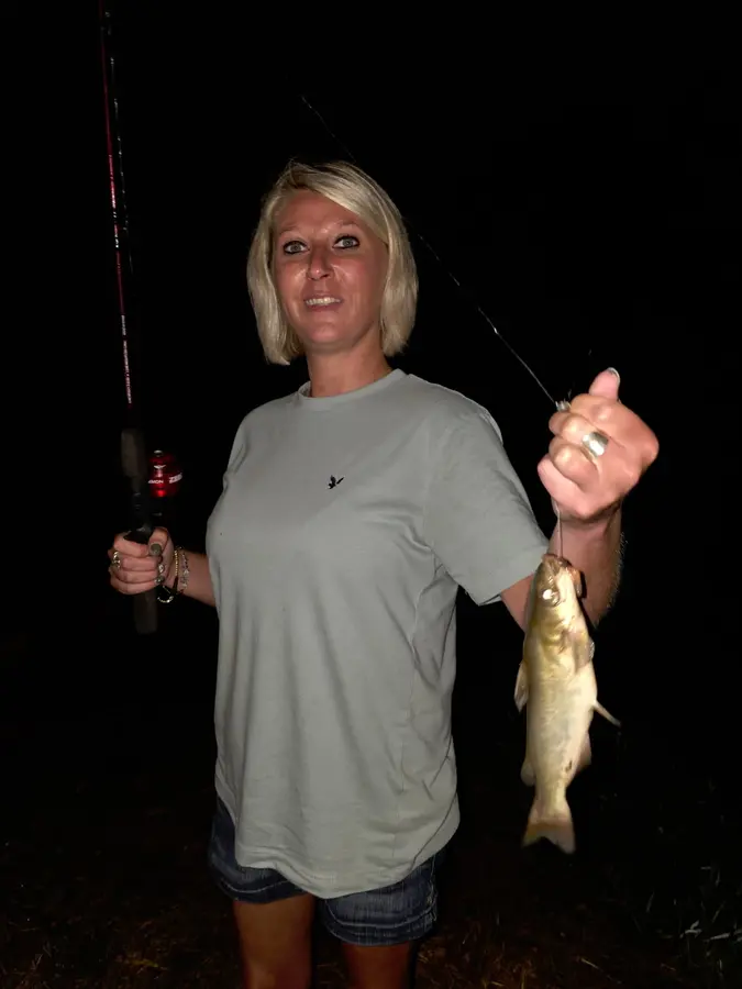 A woman outdoors at night holding a fish and a fishing rod, sharing great fishing and fun at Hillbilly Campground on Yadkin River.