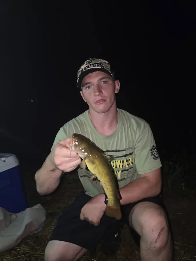 A young man seated outdoors at night and holding a fish, sharing great fishing and fun at Hillbilly Campground on Yadkin River.