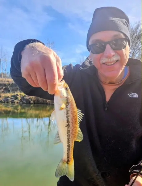 A smiling camper in sunglasses and a black knit cap holding a small bass caught from Fishing Creek.